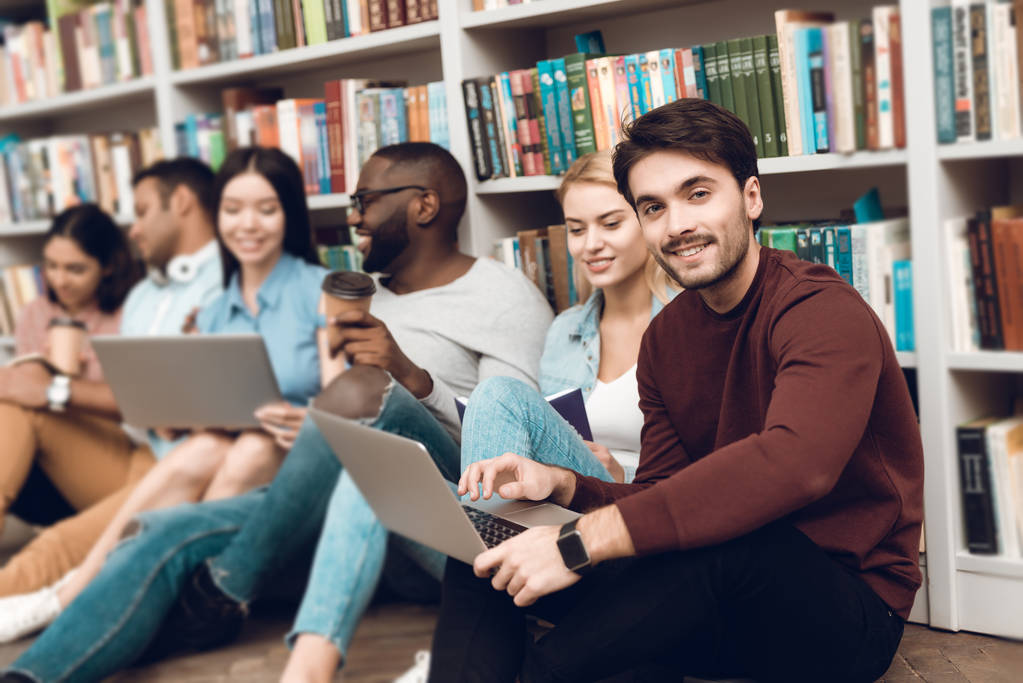Группа этнических мультикультурных студентов сидит, улыбается и разговаривает возле книжных полок в библиотеке
 - Фото, изображение