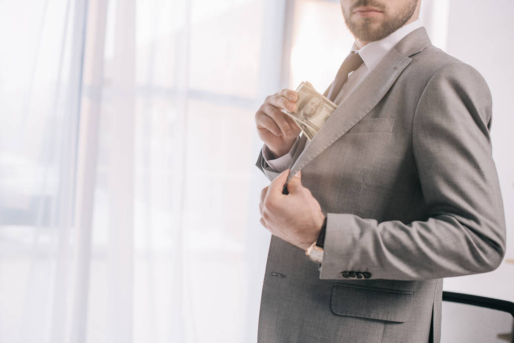 recortado tiro de hombre de negocios en traje poner billetes de dólar en el bolsillo en la oficina
 - Foto, Imagen