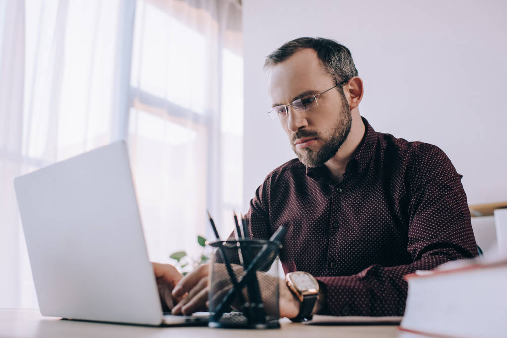 ориентированный бизнесмен в очках, работающий на ноутбуке на рабочем месте в офисе
 - Фото, изображение