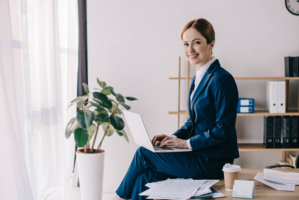 vista lateral de mulher de negócios sorridente com laptop sentado na mesa no escritório
 - Foto, Imagem