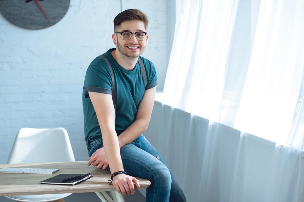 komea nuori mies silmälaseissa hymyilee kameralle istuessaan pöydällä kotitoimistossa
 - Valokuva, kuva