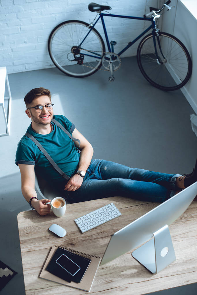 high angle view of handsome young freelancer smiling at camera while sitting at workplace  - Photo, Image