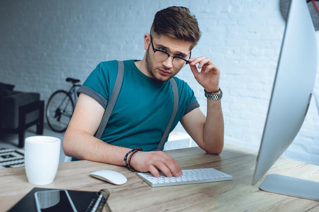 guapo joven freelancer ajustando gafas y mirando a la cámara mientras trabaja con computadora de escritorio
 - Foto, imagen