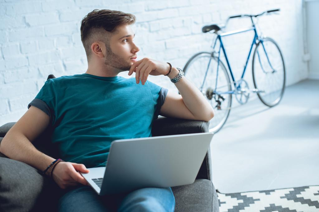 komea nuori mies käyttää kannettavaa tietokonetta ja katselee pois, kun istuu kotona
 - Valokuva, kuva