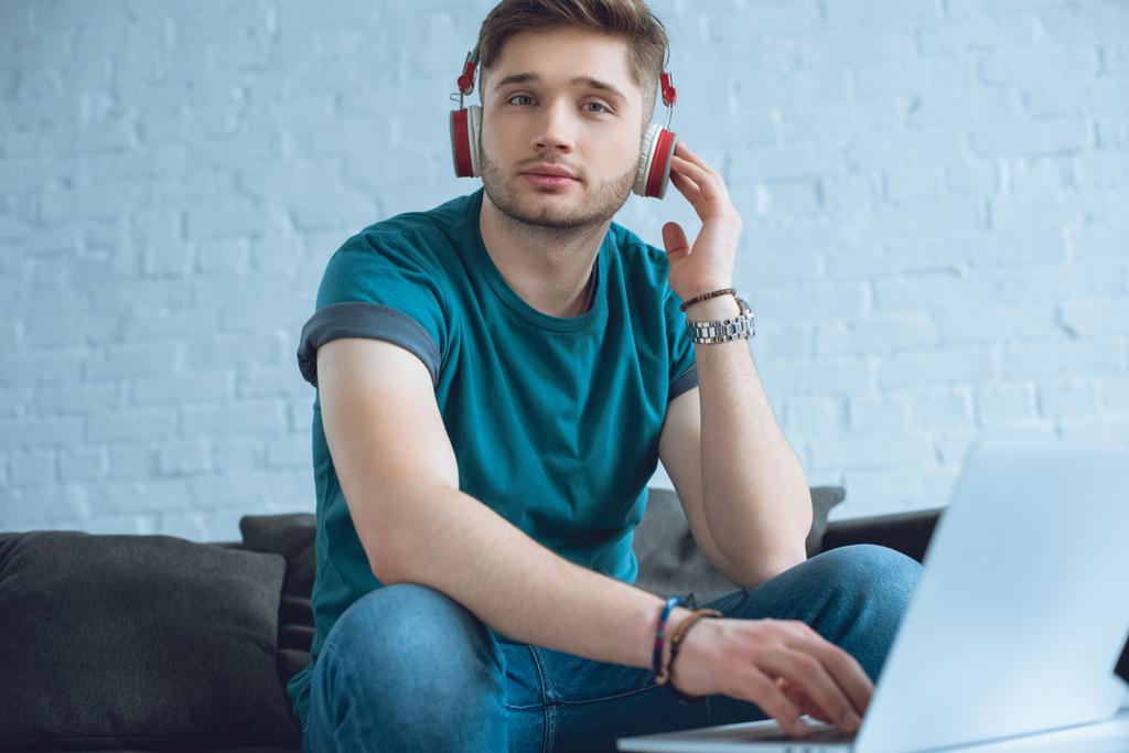 hombre joven en los auriculares usando el ordenador portátil y mirando a la cámara en casa
  - Foto, Imagen