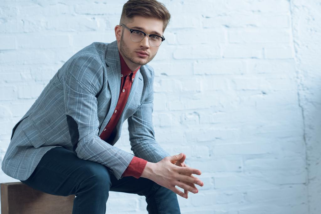 Aantrekkelijke bebaarde man pak en bril dragen en bakstenen muur zit - Foto, afbeelding