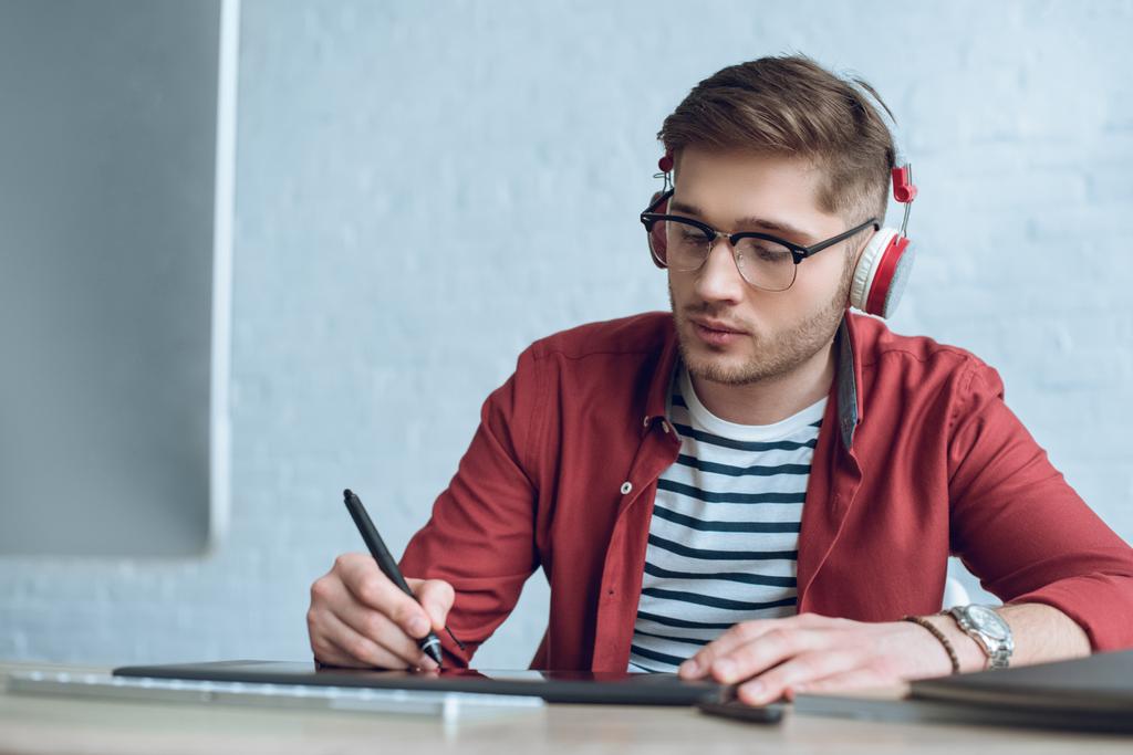 Freelancer man in headphones drawing with graphic tablet by table with computer - Photo, Image