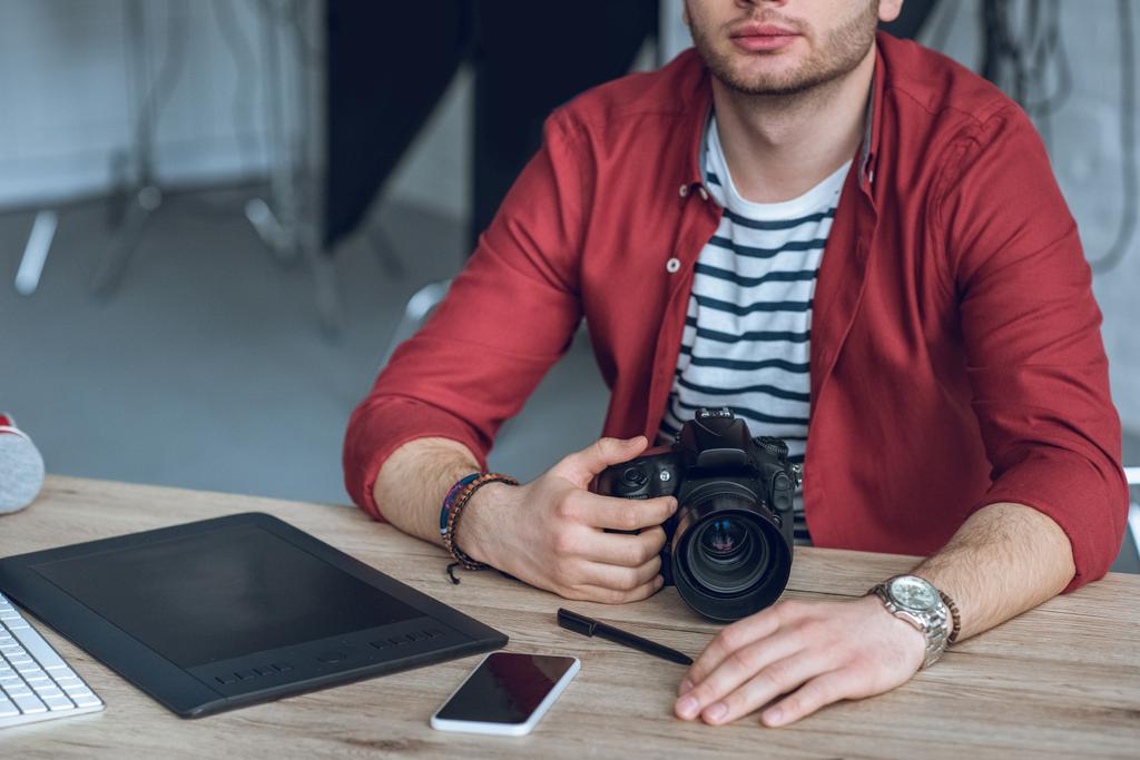Молодий бородатий чоловік тримає камеру за столом з комп'ютером
 - Фото, зображення