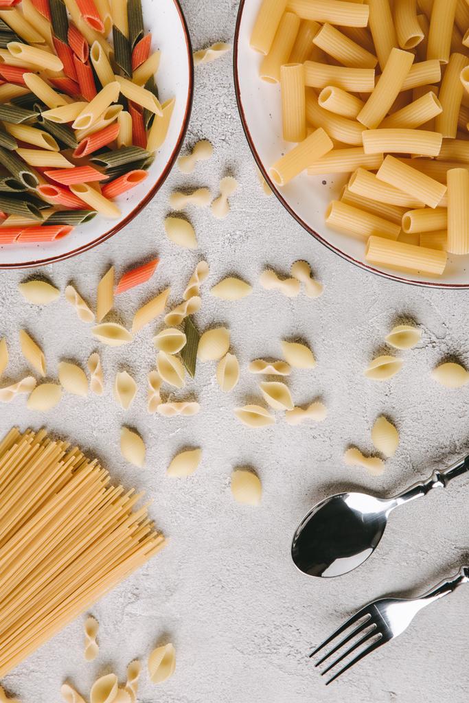 bovenaanzicht van verschillende rauwe pasta en bestek op rommelig betonnen tafelblad - Foto, afbeelding