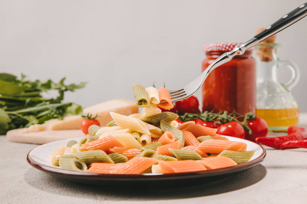 tenedor con pasta colorida perforada por encima del plato en la mesa de hormigón con varios alimentos en el fondo
 - Foto, imagen