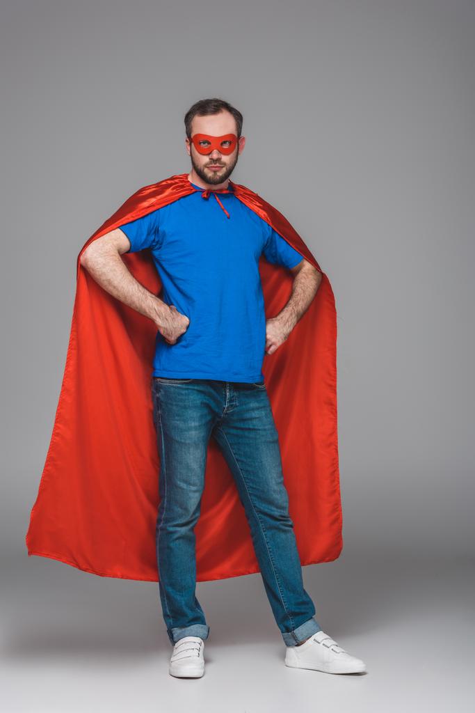 hombre de capa roja y máscara de pie con las manos en la cintura y mirando a la cámara en gris
 - Foto, Imagen