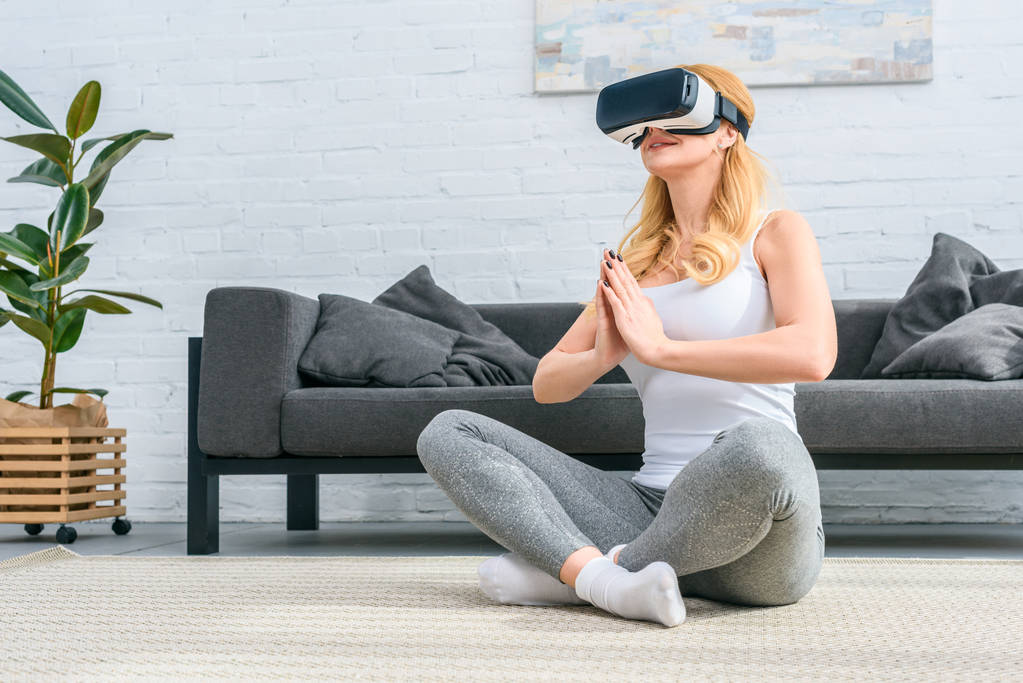 Nuori nainen harjoittelee joogaa lootusasennossa ja käyttää virtuaalitodellisuuskuulokkeita
  - Valokuva, kuva