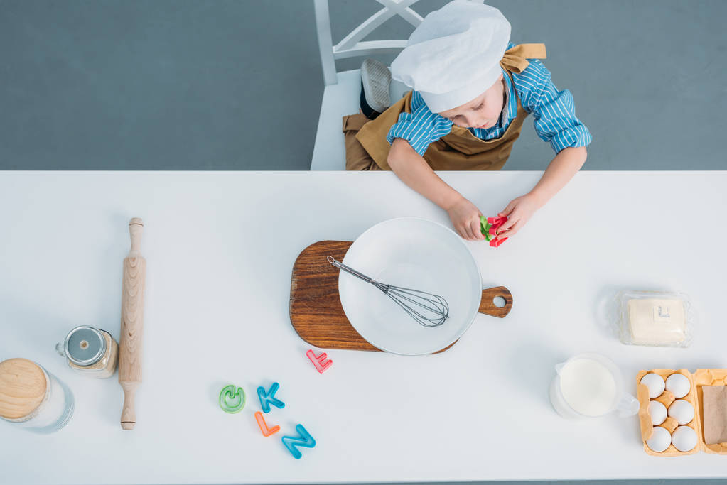 Felülnézet főzés kalap, konyhai eszközökkel és összetevőkkel asztalnál ülő kisfiú - Fotó, kép