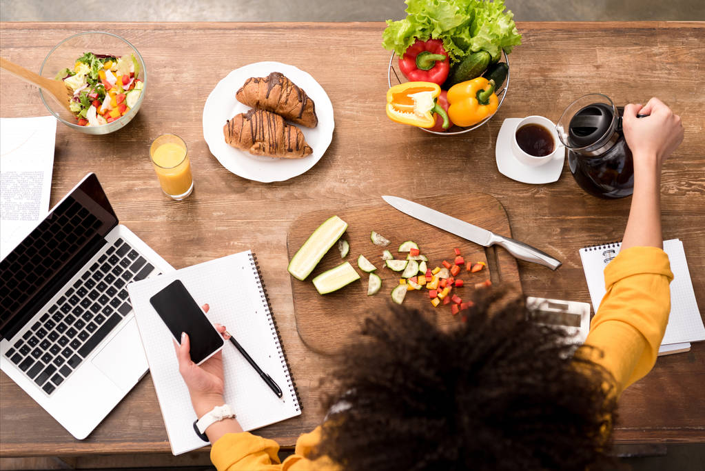 вид зверху на молоду жінку смартфон на кухні з різними продуктами харчування і ноутбуком на столі і заливання кави в чашку
 - Фото, зображення