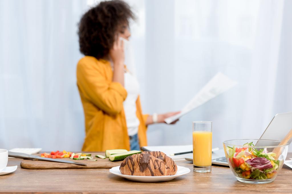 mujer joven hablando por teléfono y leyendo documentos con comida en la mesa en primer plano
 - Foto, Imagen