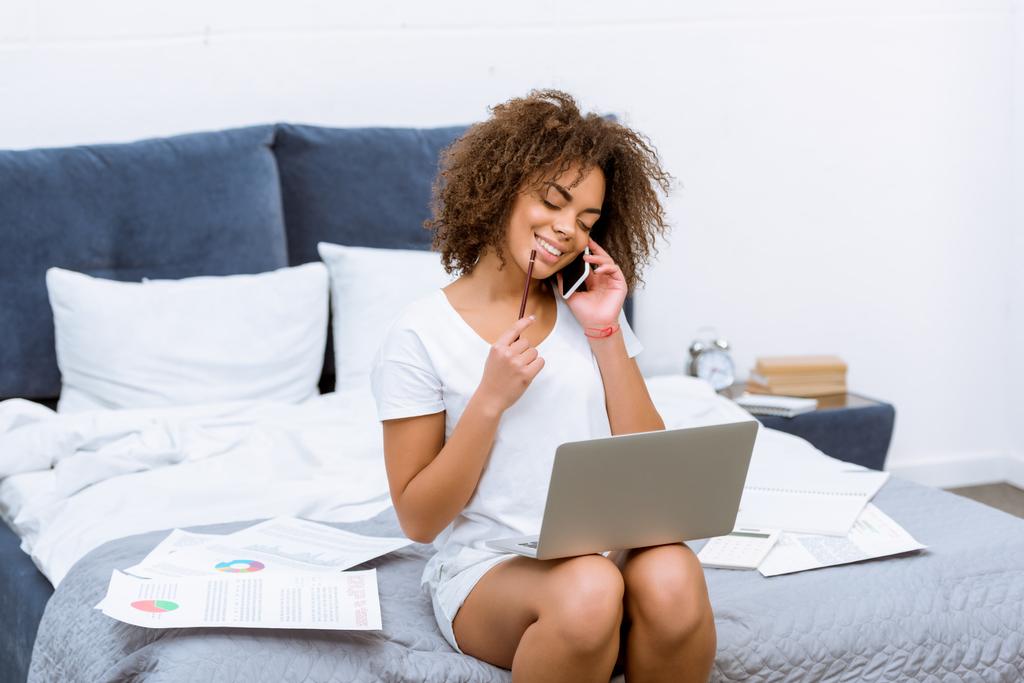 boldog fiatal nő dolgozik a laptop, és beszélt telefonon ágyban otthon - Fotó, kép