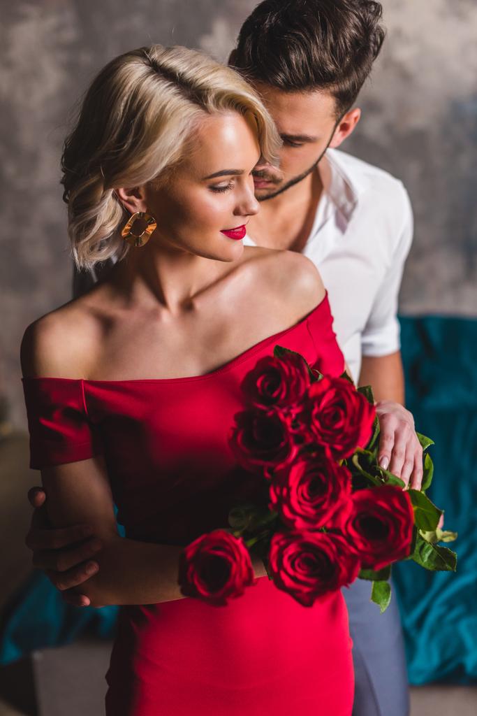 όμορφος άνδρας αγκαλιάζει χαμογελώντας αισθησιακής γυναίκας με κόκκινο φόρεμα που κατέχει όμορφα τριαντάφυλλα - Φωτογραφία, εικόνα