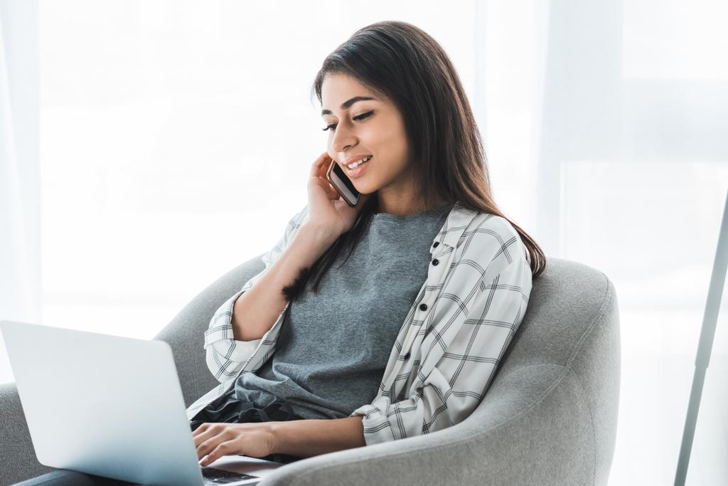 Młoda kobieta pracuje na laptopie siedząc w fotelu i rozmowy na telefon  - Zdjęcie, obraz