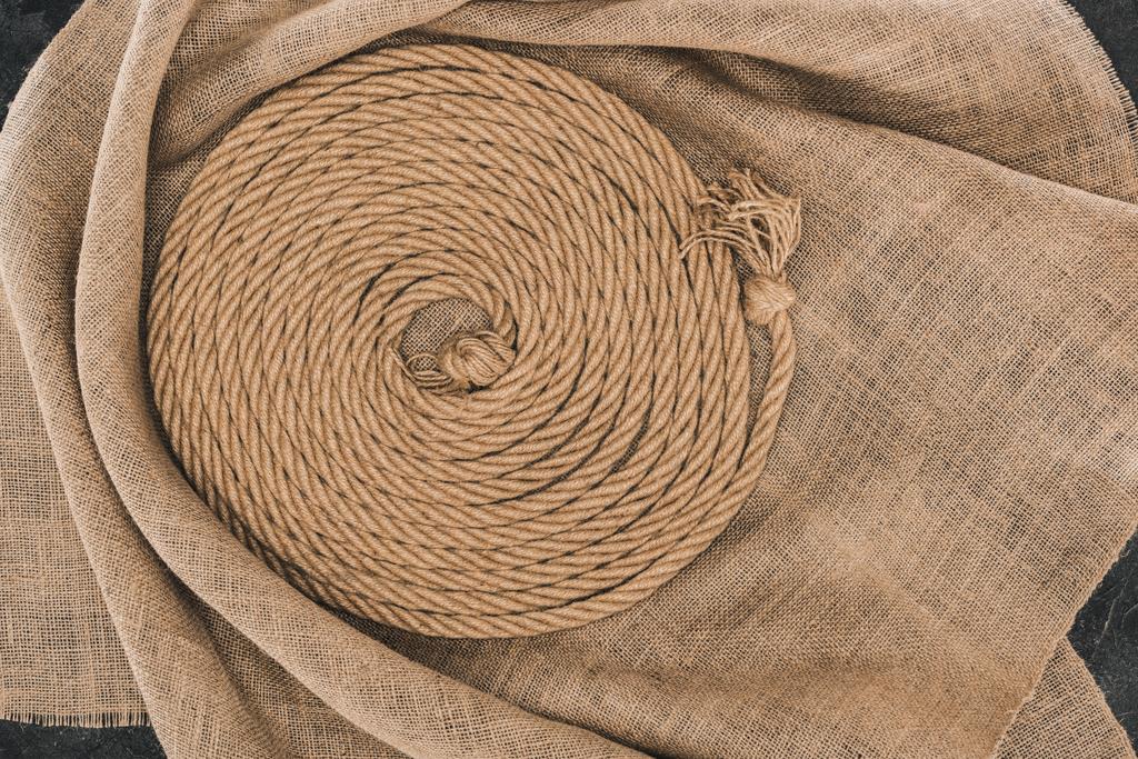 верхний вид коричневой морской верёвки, расположенной по кругу на мешковине на темной бетонной поверхности
 - Фото, изображение