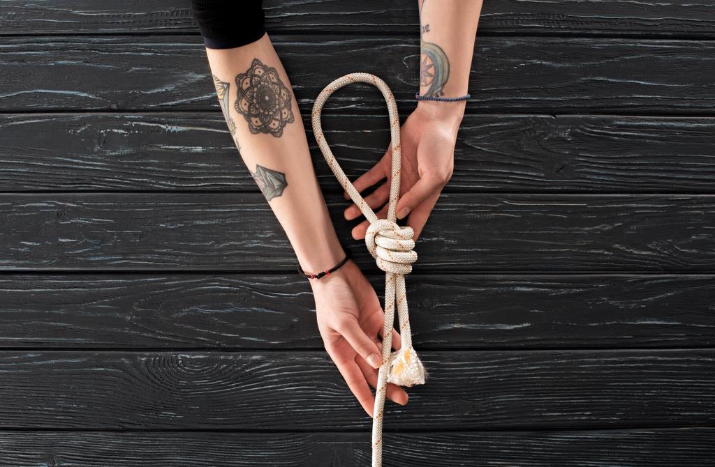 Μερική άποψη της γυναικεία τατουάζ χέρια με ναυτικά σχοινί με κόμπο σε σκούρα ξύλινη επιφάνεια - Φωτογραφία, εικόνα