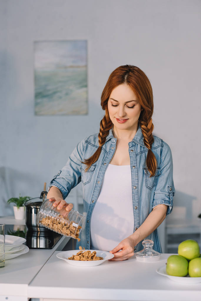 привлекательная беременная женщина выливает кукурузные хлопья в тарелку на кухне
 - Фото, изображение
