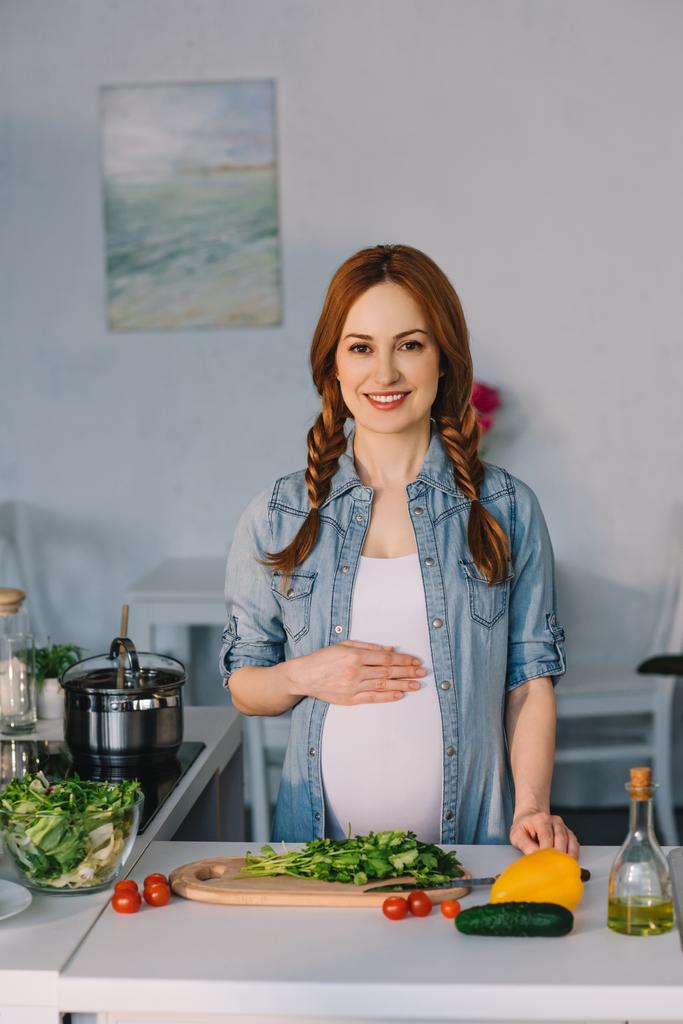 приваблива вагітна жінка торкається живота біля кухонної стійки з овочами для салату
 - Фото, зображення