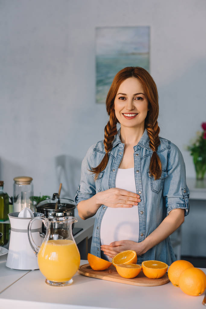souriant femme enceinte toucher le ventre et regarder la caméra à la cuisine
 - Photo, image