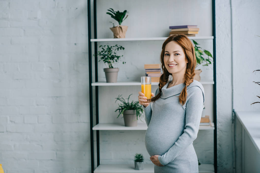 atractiva mujer embarazada sonriente sosteniendo un vaso de jugo de naranja y mirando a la cámara en casa
 - Foto, Imagen