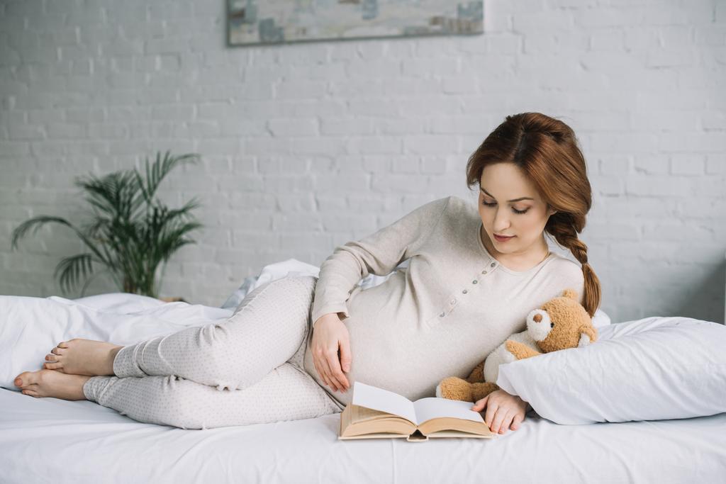 attractive pregnant woman lying on bed with teddy and reading book in bedroom - Photo, Image