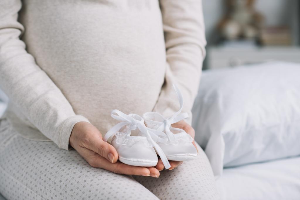 imagem cortada de mulher grávida segurando sapatos recém-nascidos no quarto
 - Foto, Imagem