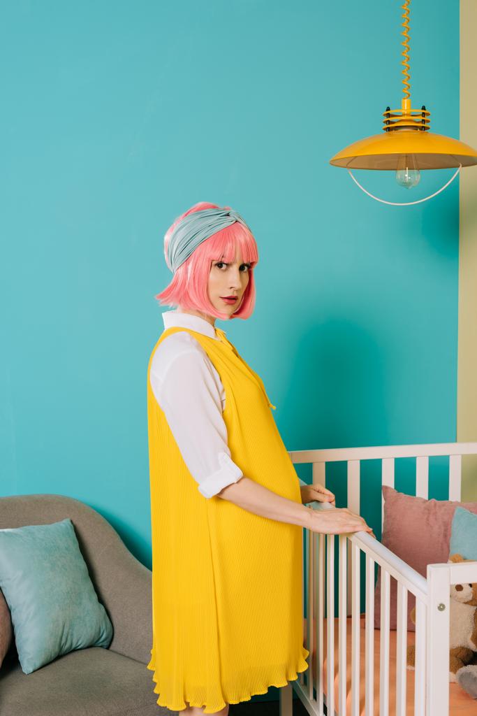 stylish pregnant pin up woman with pink hair standing near baby cot in child room - Photo, Image