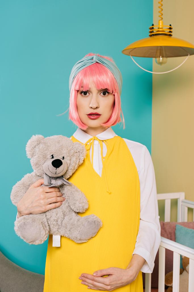 πορτρέτο του ρετρό στυλ pin έγκυος γυναίκα με ροζ μαλλιά κρατώντας αρκουδάκι στο παιδικό δωμάτιο - Φωτογραφία, εικόνα