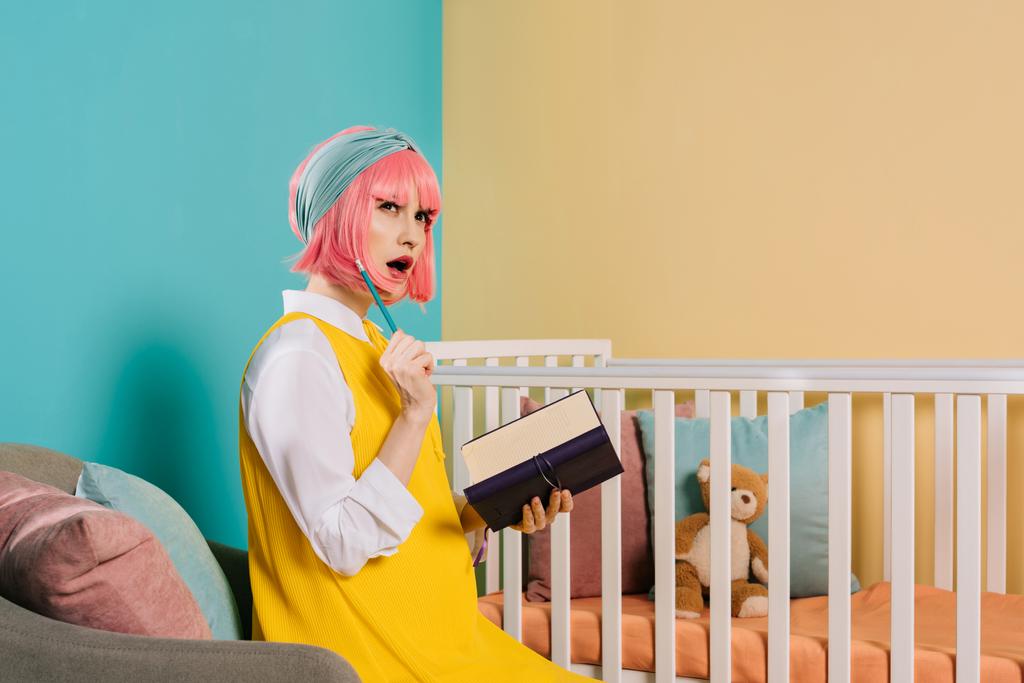 verbaasd stijlvolle zwangere pin-up vrouw met roze haar bedrijf notitieboekje en potlood in de buurt van babybedje - Foto, afbeelding