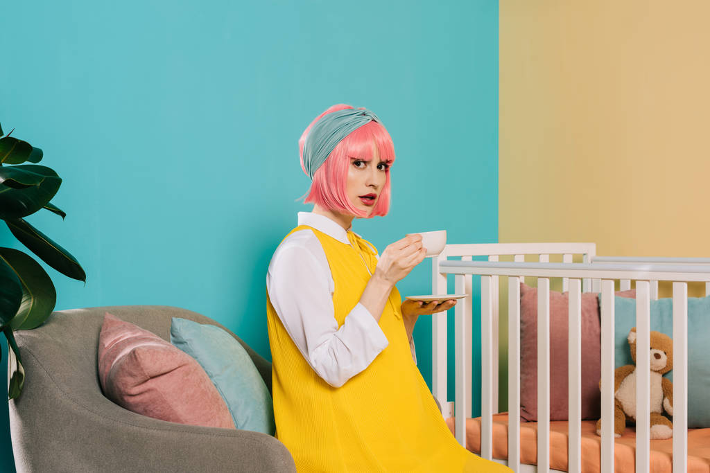retro estilo grávida pin up mulher com cabelo rosa sentado perto berço com xícara de chá e olhando para a câmera
 - Foto, Imagem