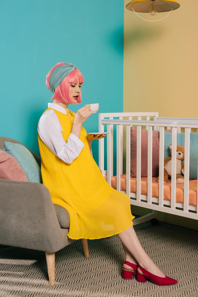 Schwangere Pin-up-Frau im Retro-Stil mit rosa Haaren trinkt Tee in der Nähe des Babybettes im Kinderzimmer - Foto, Bild