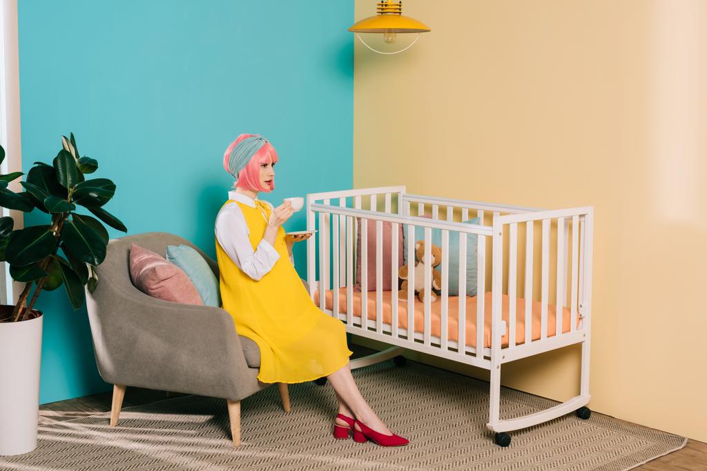 Widok z boku retro stylu PIN w ciąży kobieta siedzi w pobliżu łóżeczko z kubkiem herbaty w pokoju dziecka - Zdjęcie, obraz