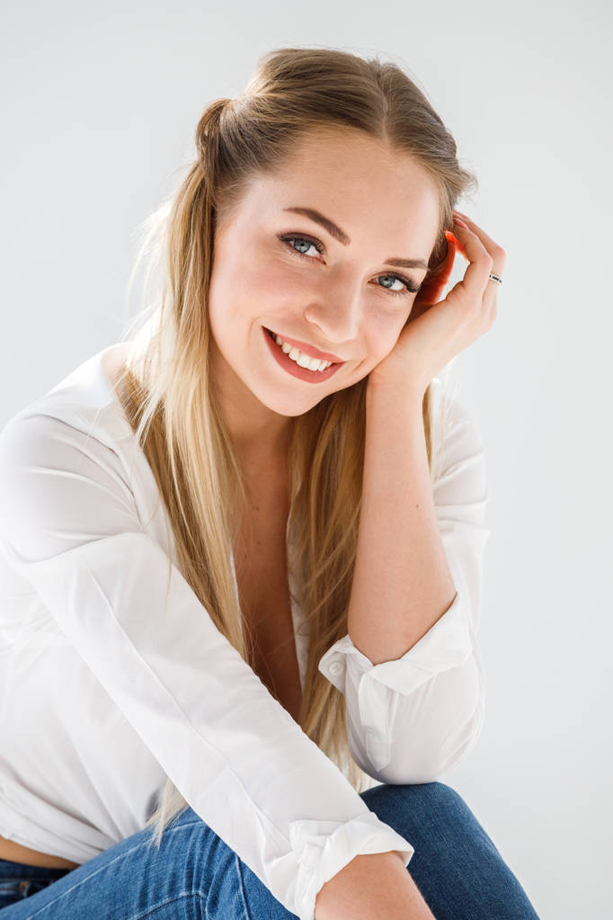 Ritratto di ragazza bionda sexy che indossa jeans blu e camicia bianca seduta su sfondo bianco dello studio
 - Foto, immagini