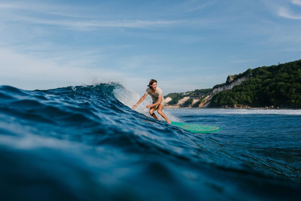 Вид сбоку человека в мокрой футболке, катающегося на синих океанских волнах на доске для серфинга
 - Фото, изображение