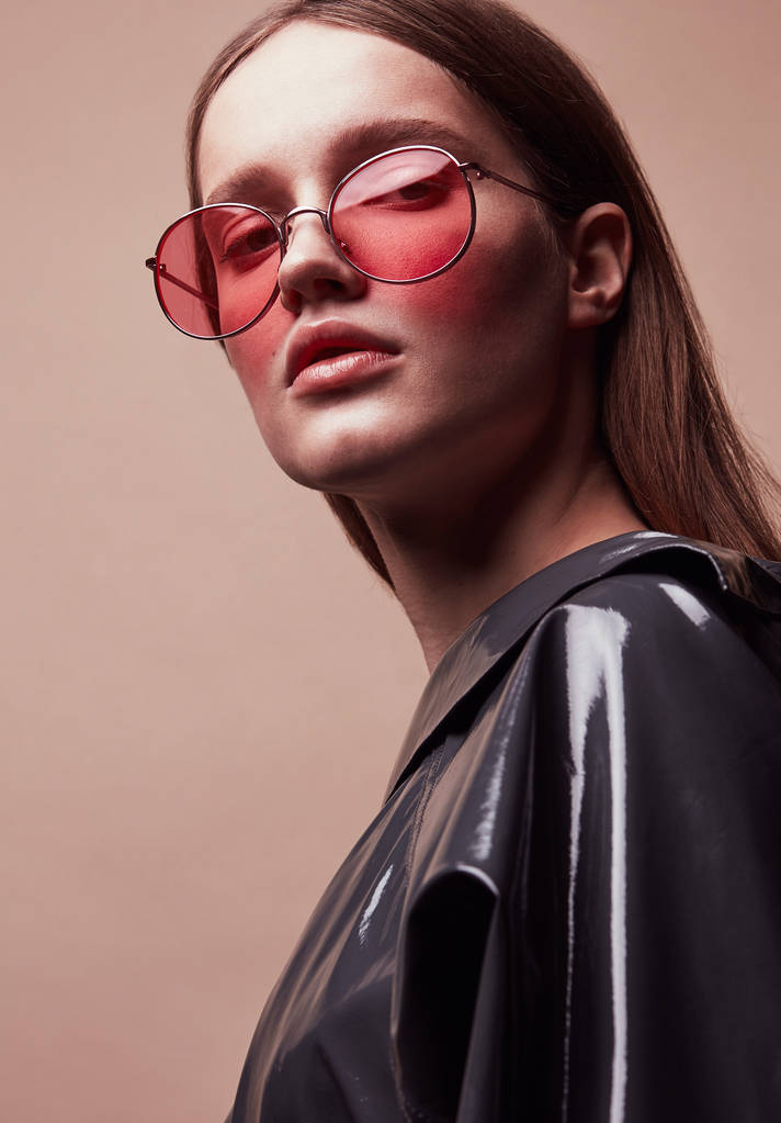Güzel genç kadın moda portre yuvarlak kırmızı güneş gözlüğü ve stüdyoda poz gri lateks ceket - Fotoğraf, Görsel