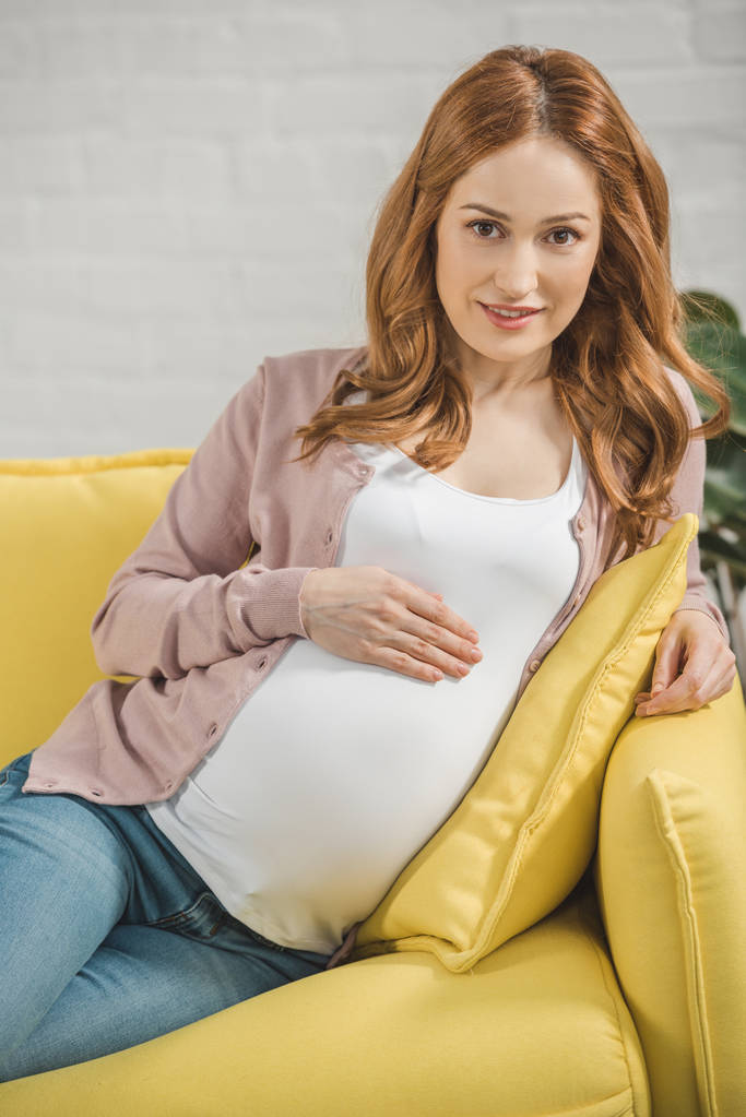 hermosa mujer embarazada tocando el vientre y sonriendo a la cámara en casa
 - Foto, Imagen