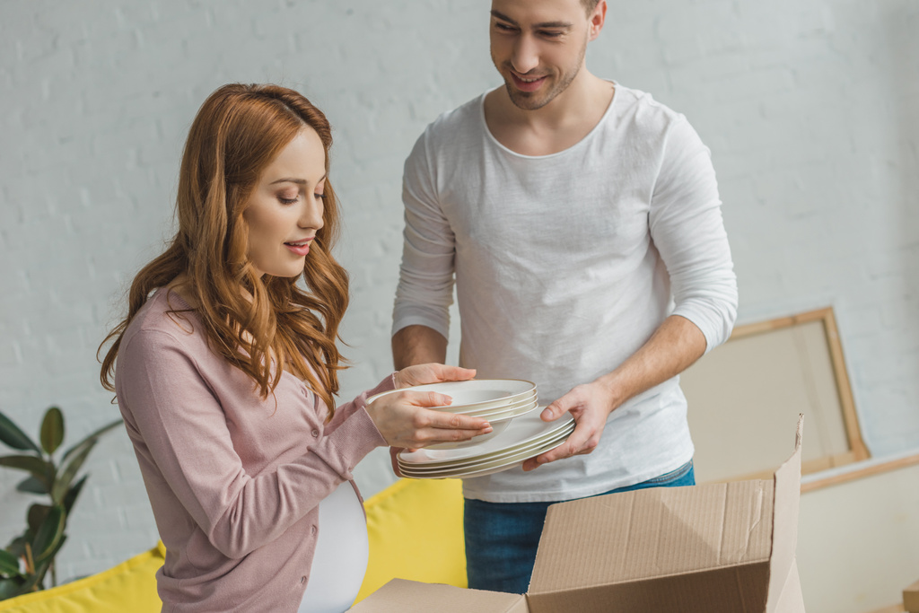 young pregnant couple packing plates in cardboard box during relocation - Photo, Image