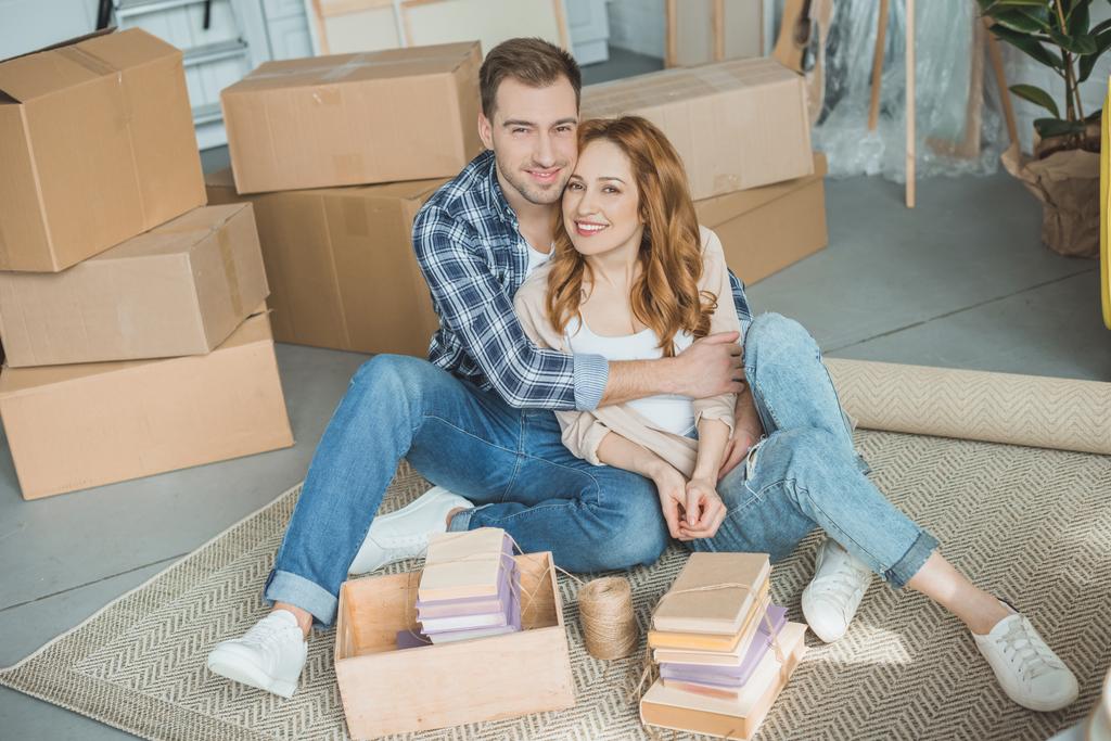 счастливая молодая пара, сидящая вместе и улыбающаяся в камеру, упаковывая коробки во время переезда
 - Фото, изображение