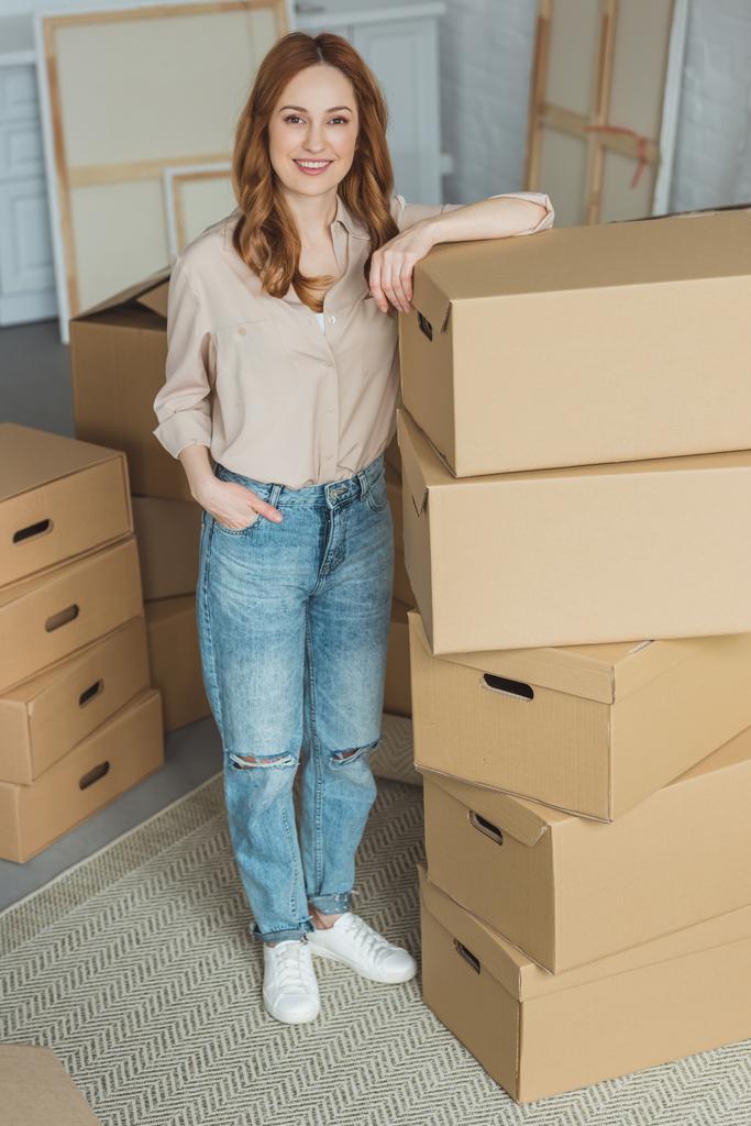 mulher sorridente atraente inclinando-se em caixas de papelão no novo apartamento, conceito de realocação
 - Foto, Imagem