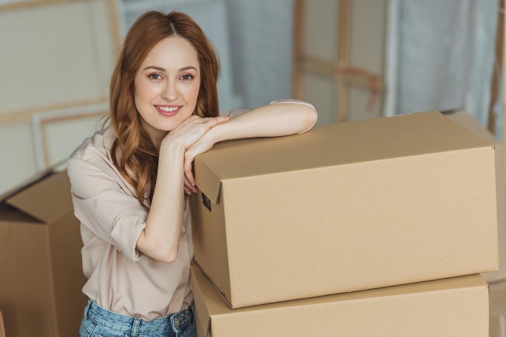 ελκυστική γυναίκα χαμογελαστή κλίνει σε κουτιά από χαρτόνι στο νέο διαμέρισμα, η έννοια της μετεγκατάστασης - Φωτογραφία, εικόνα
