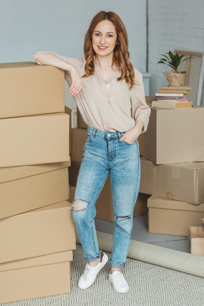 smiling woman leaning on pile of cardboard boxes at new home, relocation concept  - Photo, Image