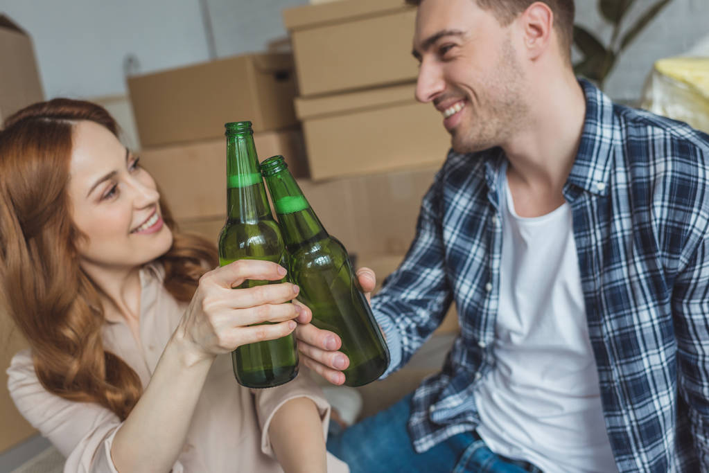young couple clinking bottles of beer at new apartment, relocation concept - Photo, Image