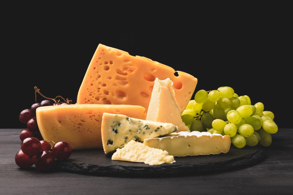 Closeup shot of different types of cheese on board with grapes on black  - Photo, Image