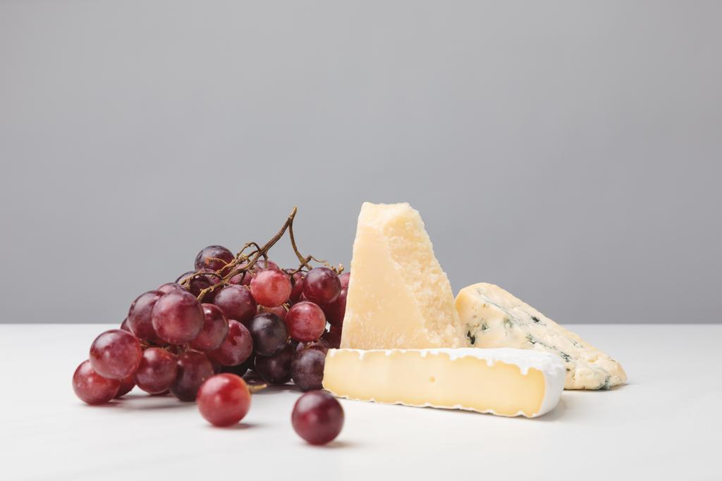 Nahaufnahme von Brie, Cheddar und Blauschimmelkäse mit Trauben auf Grau - Foto, Bild