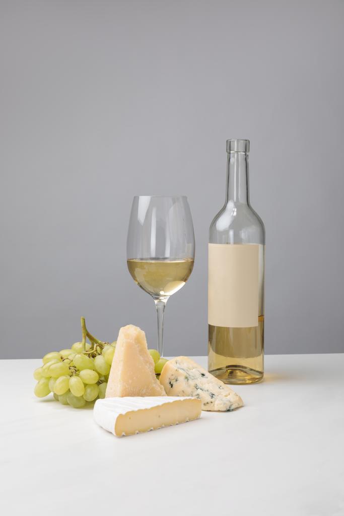 Different types of cheese, grapes, bottle and white wine glass on gray - Photo, Image