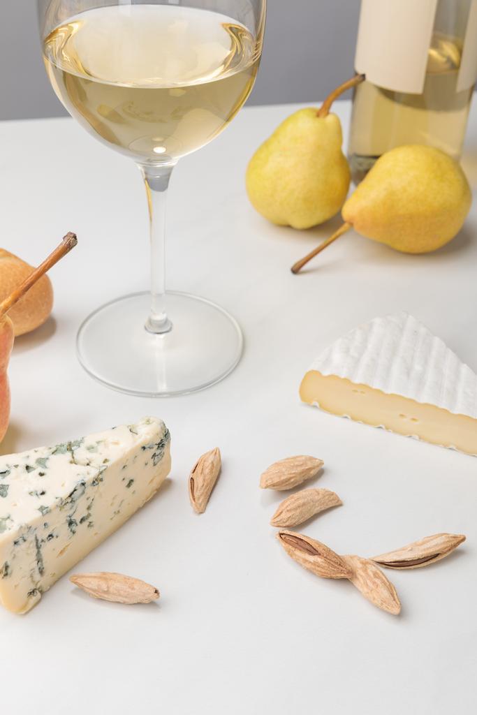 Nahaufnahme von Brie und Blauschimmelkäse mit Mandeln, Birnen, Weinglas, Flasche und Baguette auf grau  - Foto, Bild